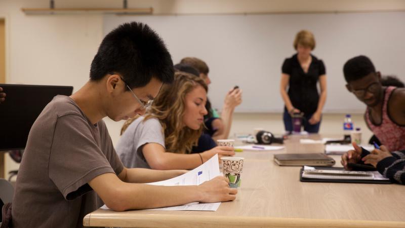 Summer Institute students reading a Workshop Handout in a Briggs Hall Classroom. Kate Zoromski, the Workshop host is standing at the end of the table.